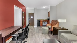 A modern living room with a wooden desk, black office chair, and a sofa. A kitchen with a sink and cabinets is visible in the background. - Best Western Plus Rose City Suites (Welland)