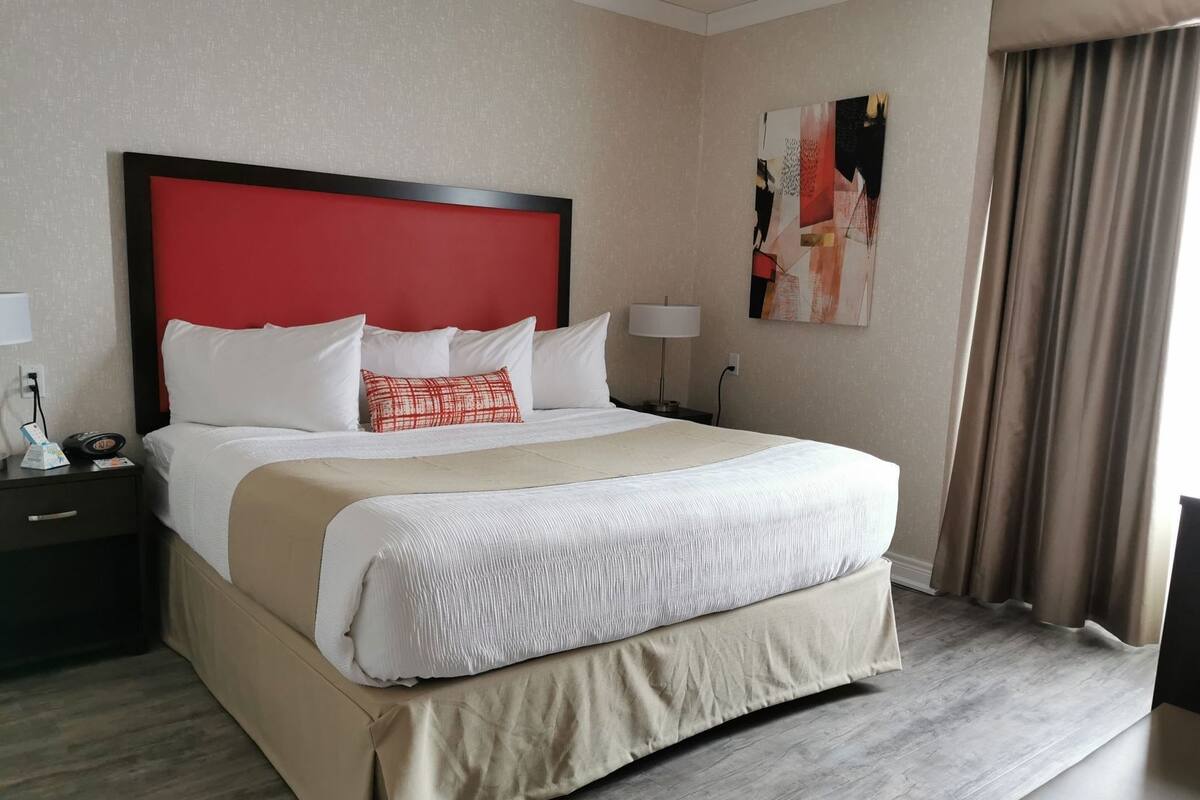 A hotel room with a large bed, a red headboard, white bedding, a beige accent, a nightstand with a lamp, and a painting on the wall.