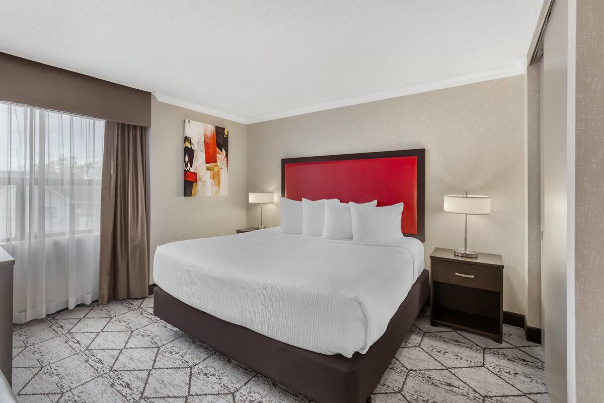 A hotel room with a large bed, a red headboard, a nightstand with a lamp, and a window with curtains.