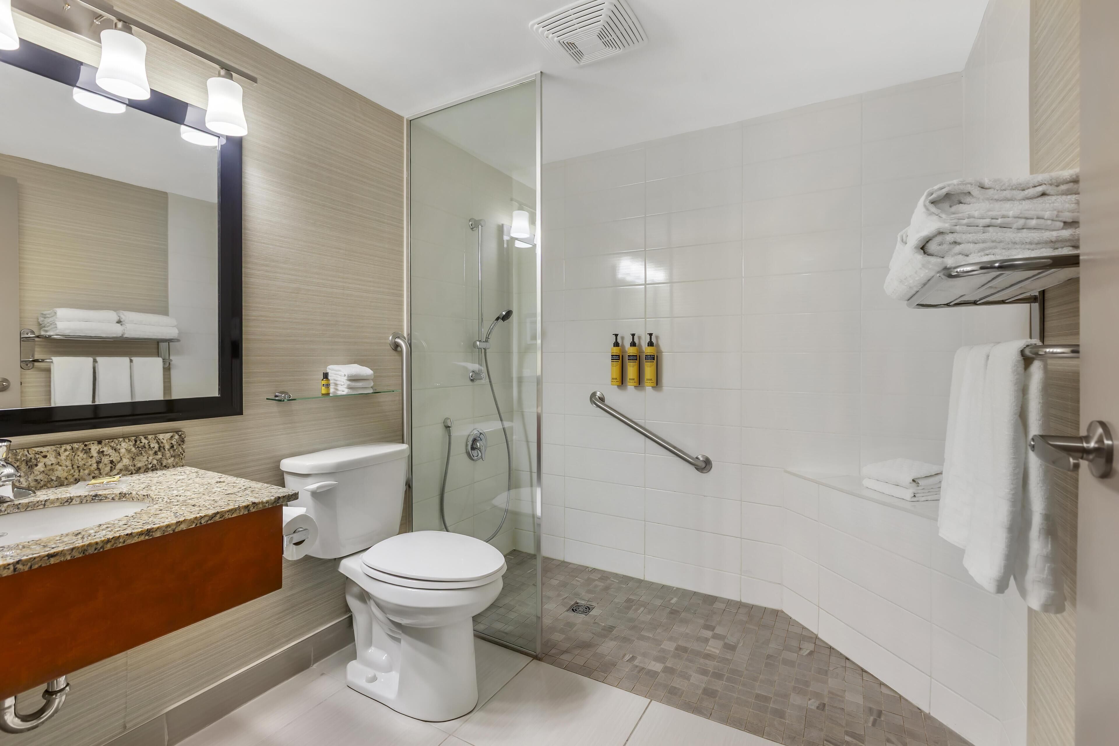 A modern bathroom with a glass-enclosed shower, a granite countertop, a toilet, and a mirror with lights.