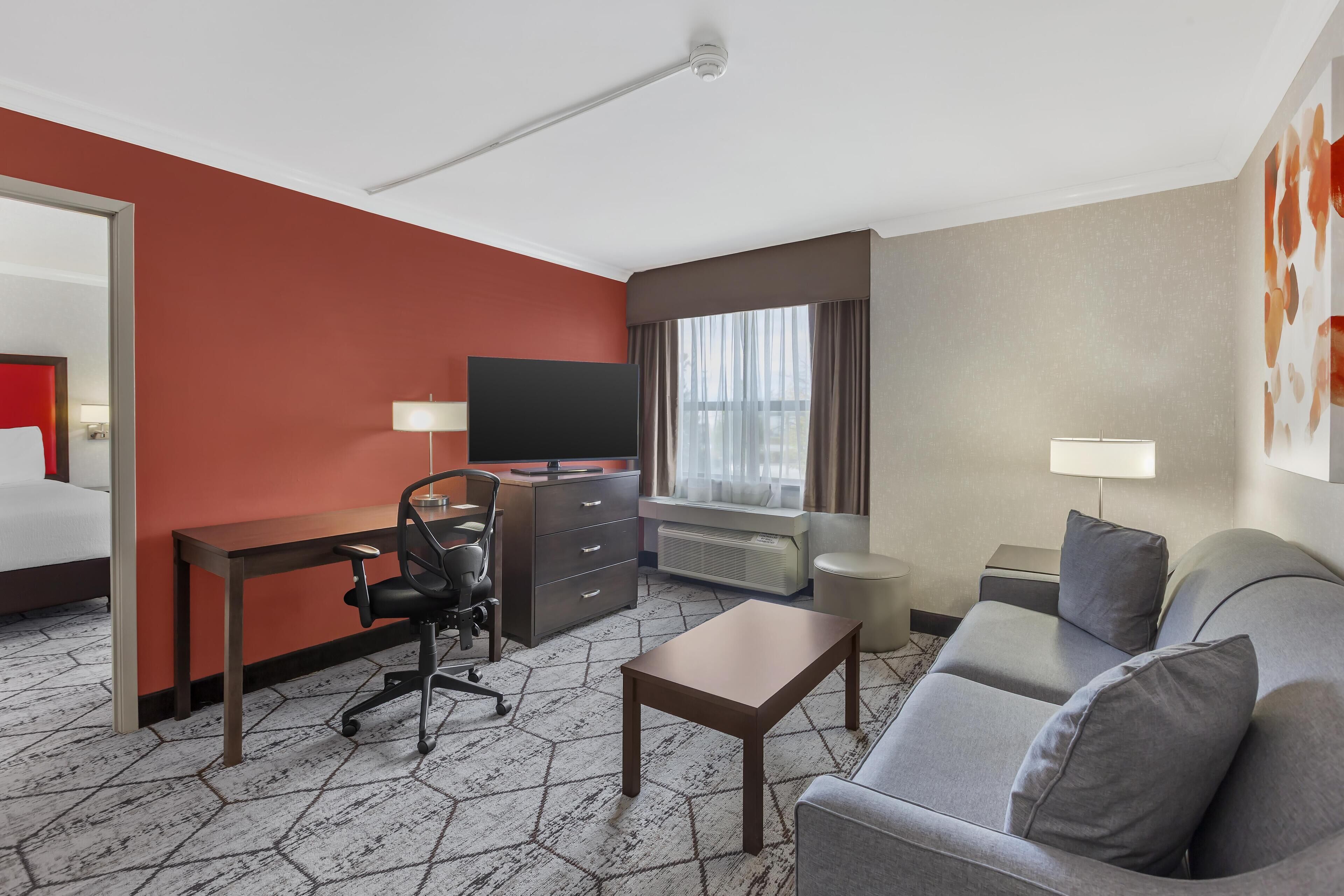 A hotel room with a red accent wall, a gray sofa, a desk with a chair, a television, and a window with curtains.