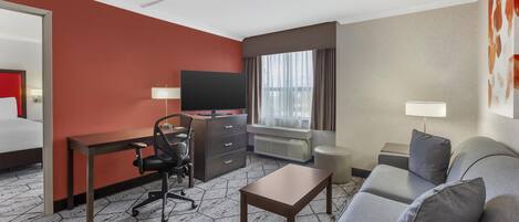 A hotel room with a red accent wall, a gray sofa, a desk with a chair, a television, and a window with curtains.