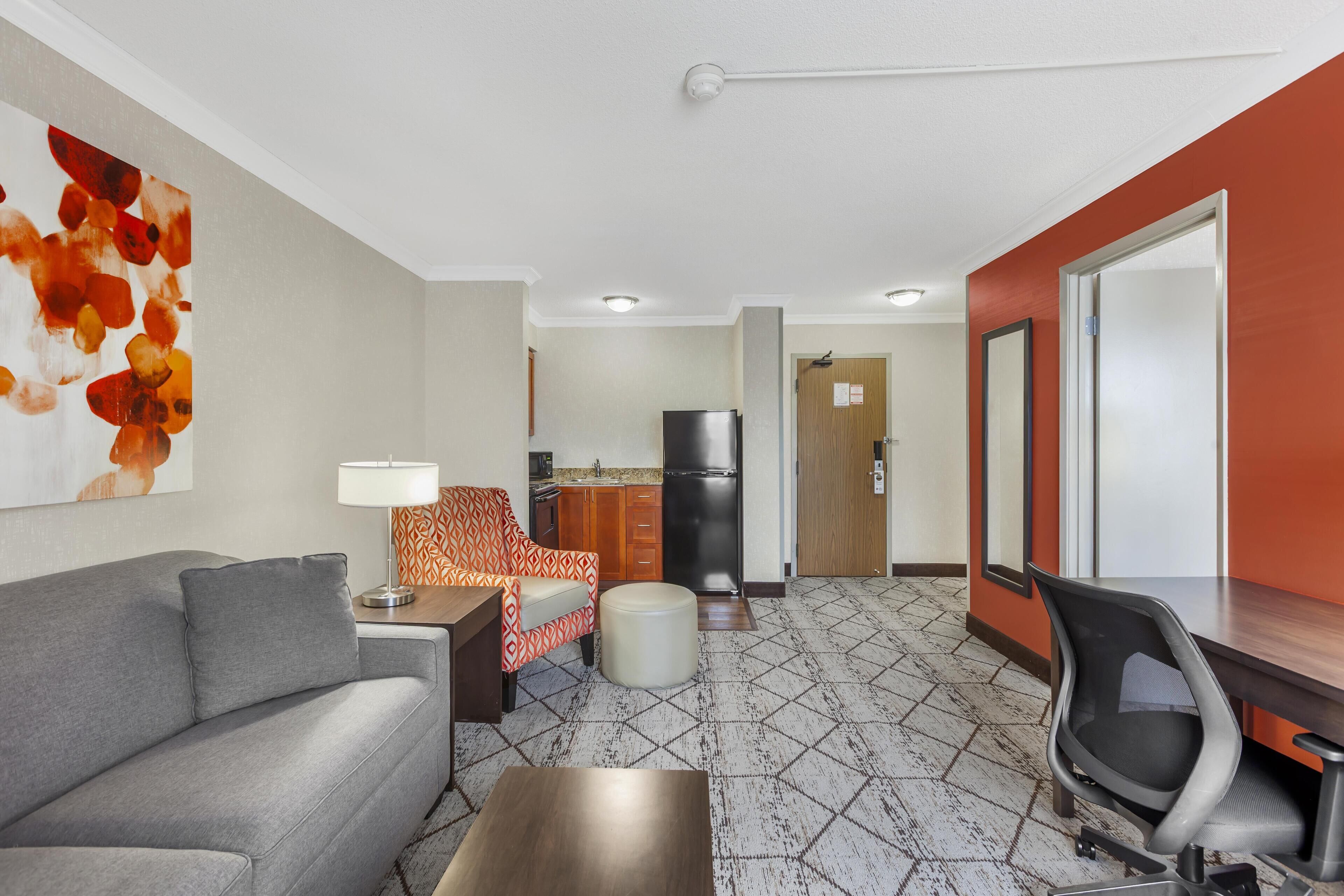 Une chambre d’hôtel moderne avec un canapé gris, un fauteuil à motifs, une petite table ronde, un bureau avec une chaise, un réfrigérateur et un coin cuisine équipé de placards et d’un évier.
