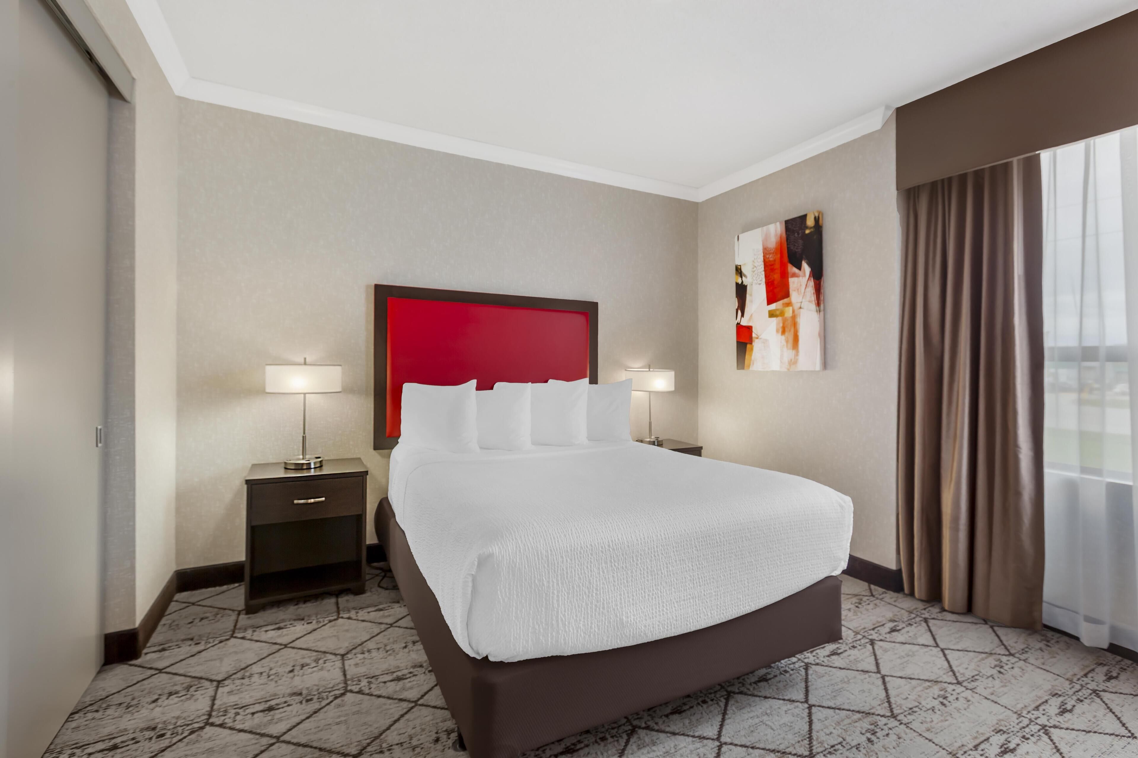 Habitación de hotel con una cama grande, dos mesitas de noche con lámparas, una pared de acento roja, un cuadro y una ventana con cortinas.