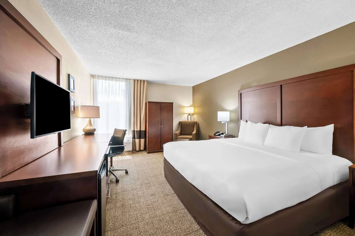 Ein Hotelzimmer mit einem großen Bett, einem Schreibtisch mit Fernseher, einem Sessel, einer Schreibtischlampe und einem Nachttisch mit Lampe.