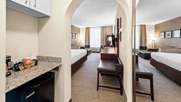 Quarto de hotel com cama, escrivaninha, cadeira, mesa pequena, cafeteira e lixeira.