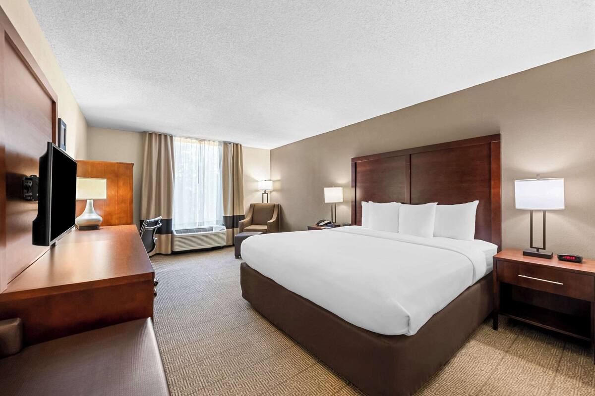 Ein Hotelzimmer mit einem großen Bett, einem Schreibtisch mit Lampe, einem Sessel und einem Fernseher.