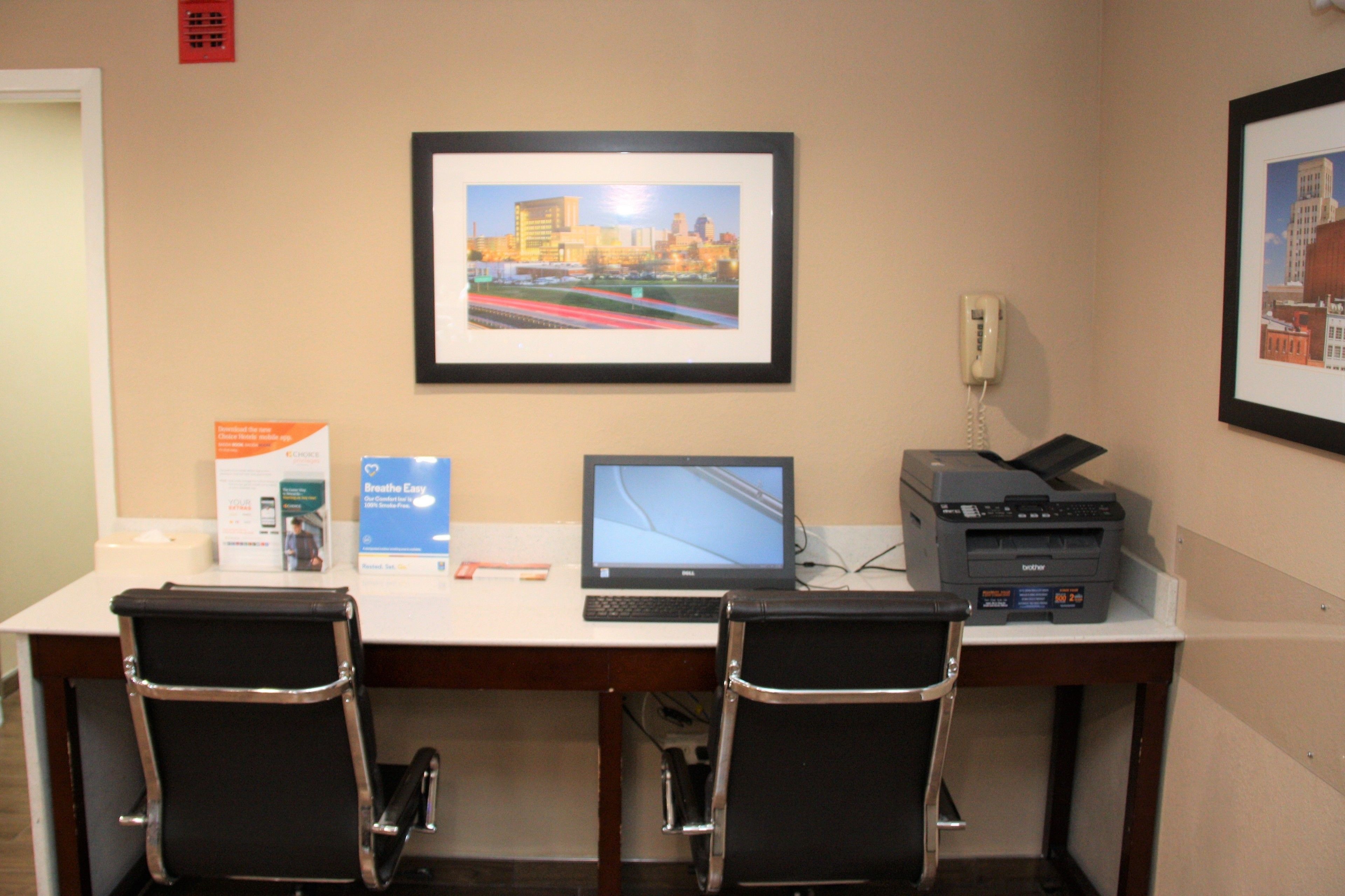 A desk with a computer, printer, and two chairs. There are framed pictures on the wall and a phone on the desk.