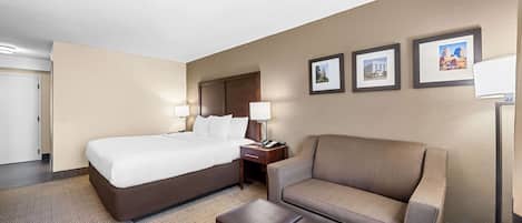 Habitación de hotel con una cama grande, un sofá, una mesita y una mesita de noche. Hay tres cuadros en la pared.