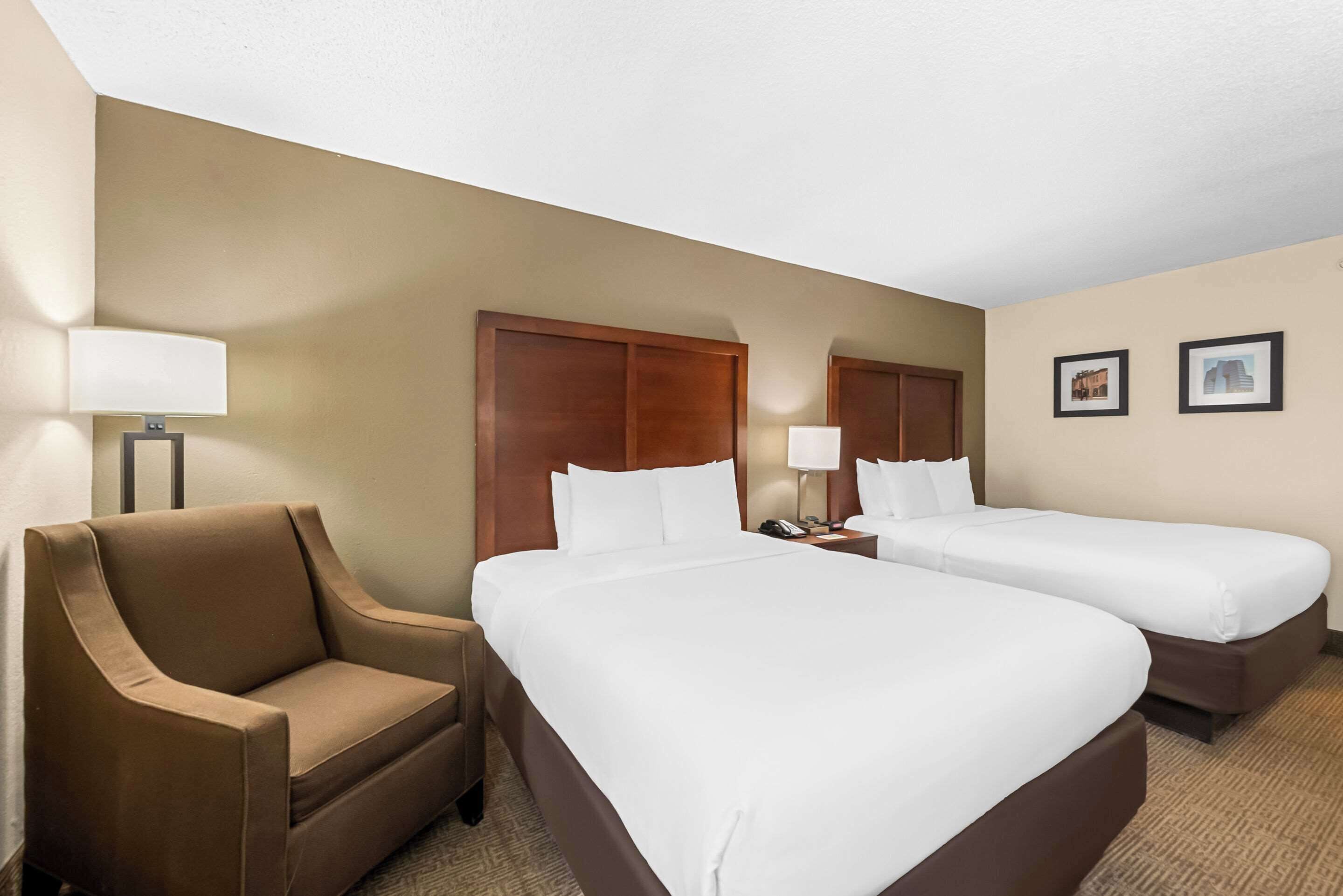 A hotel room with two single beds, a brown armchair, a nightstand with a lamp, and two framed pictures on the wall.