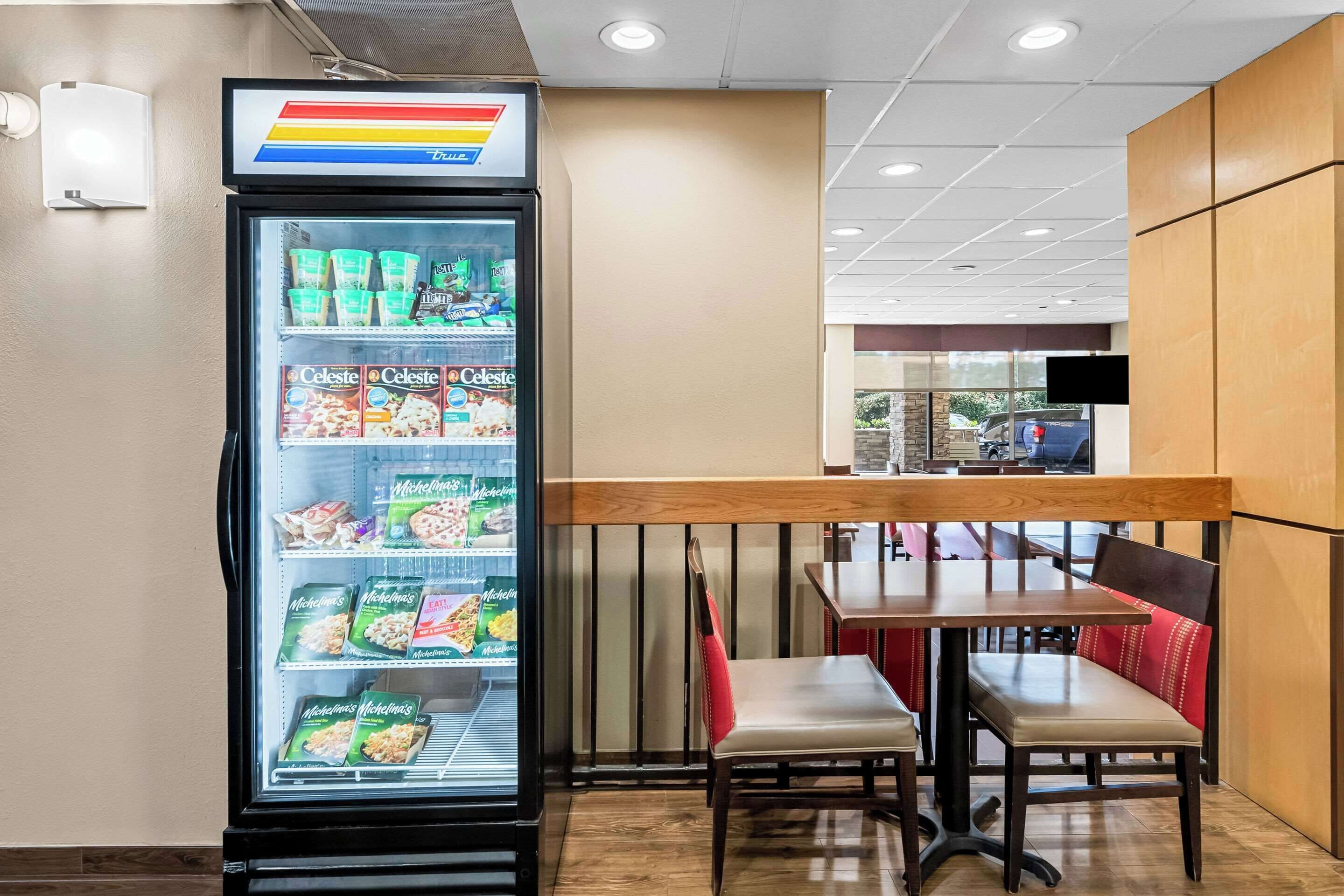 A self-service beverage dispenser with various drink options, a seating area with tables and chairs, and a view of the outside through a window.