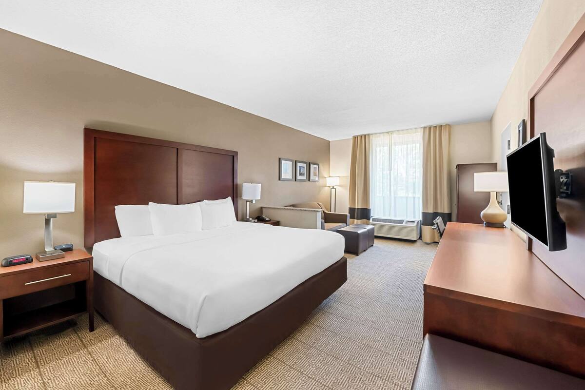 Ein Hotelzimmer mit einem großen Bett, Nachttischen, einem Schreibtisch mit Stuhl, einem Fernseher und einem Fenster mit Vorhängen.