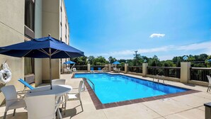 Piscina com espreguiçadeiras e guarda-sóis, emoldurada por um edifício sob um céu claro.