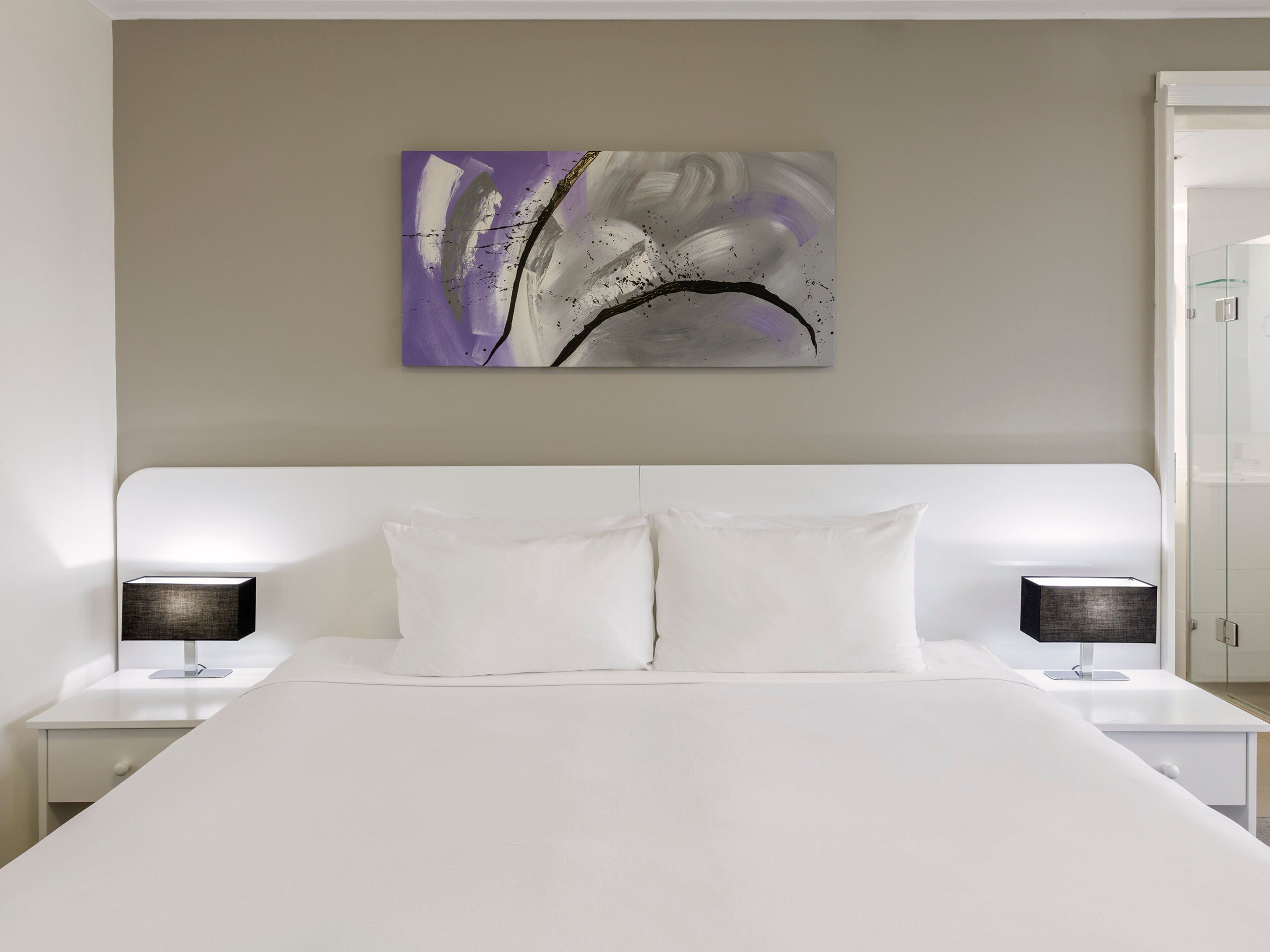 Habitación de hotel moderna con una cama grande, cabecera blanca, dos mesitas de noche con lámparas y un cuadro abstracto colgado en la pared.