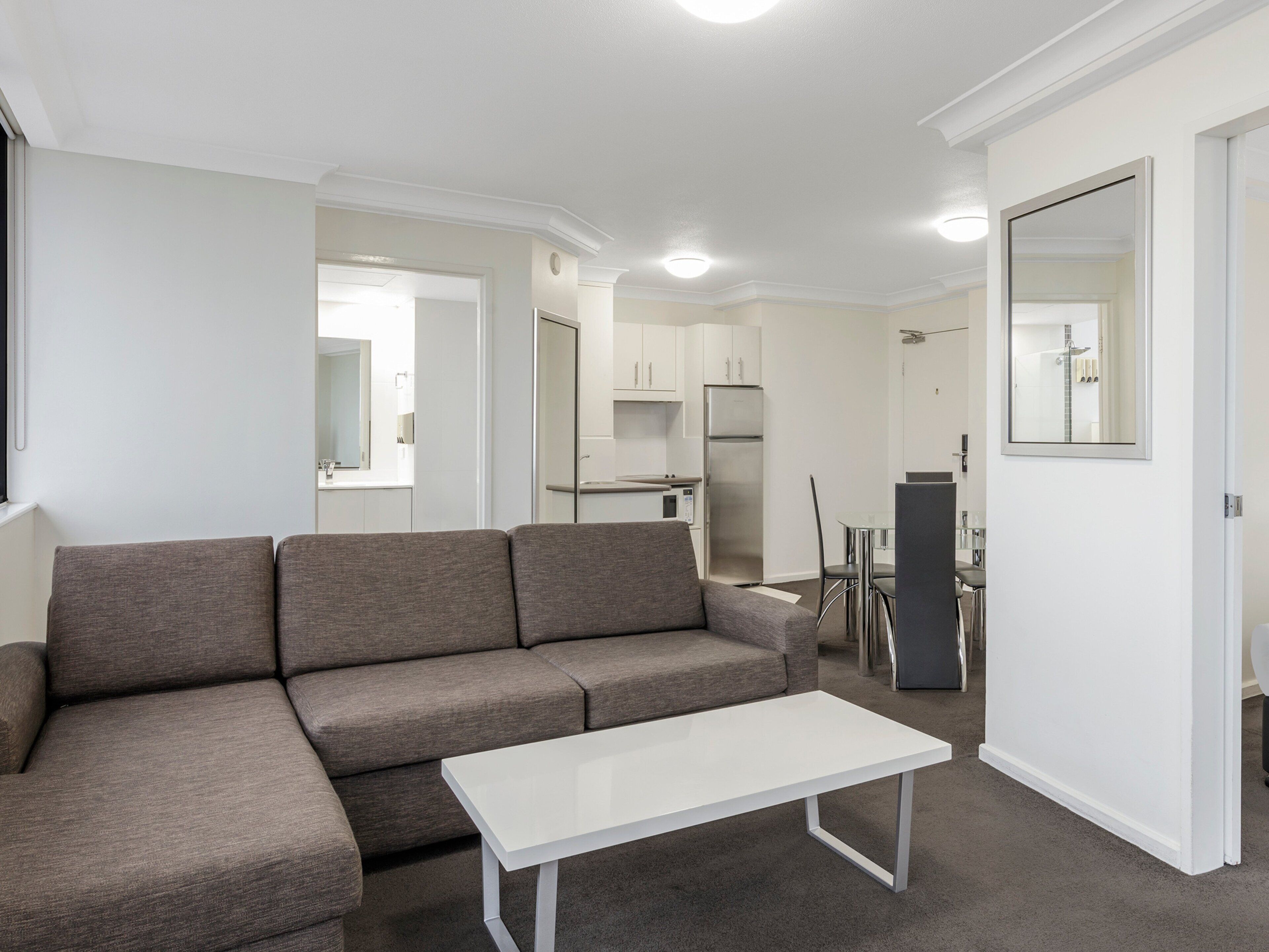 Una sala de estar moderna con un sofá gris, una mesa de centro blanca y una zona de cocina al fondo.
