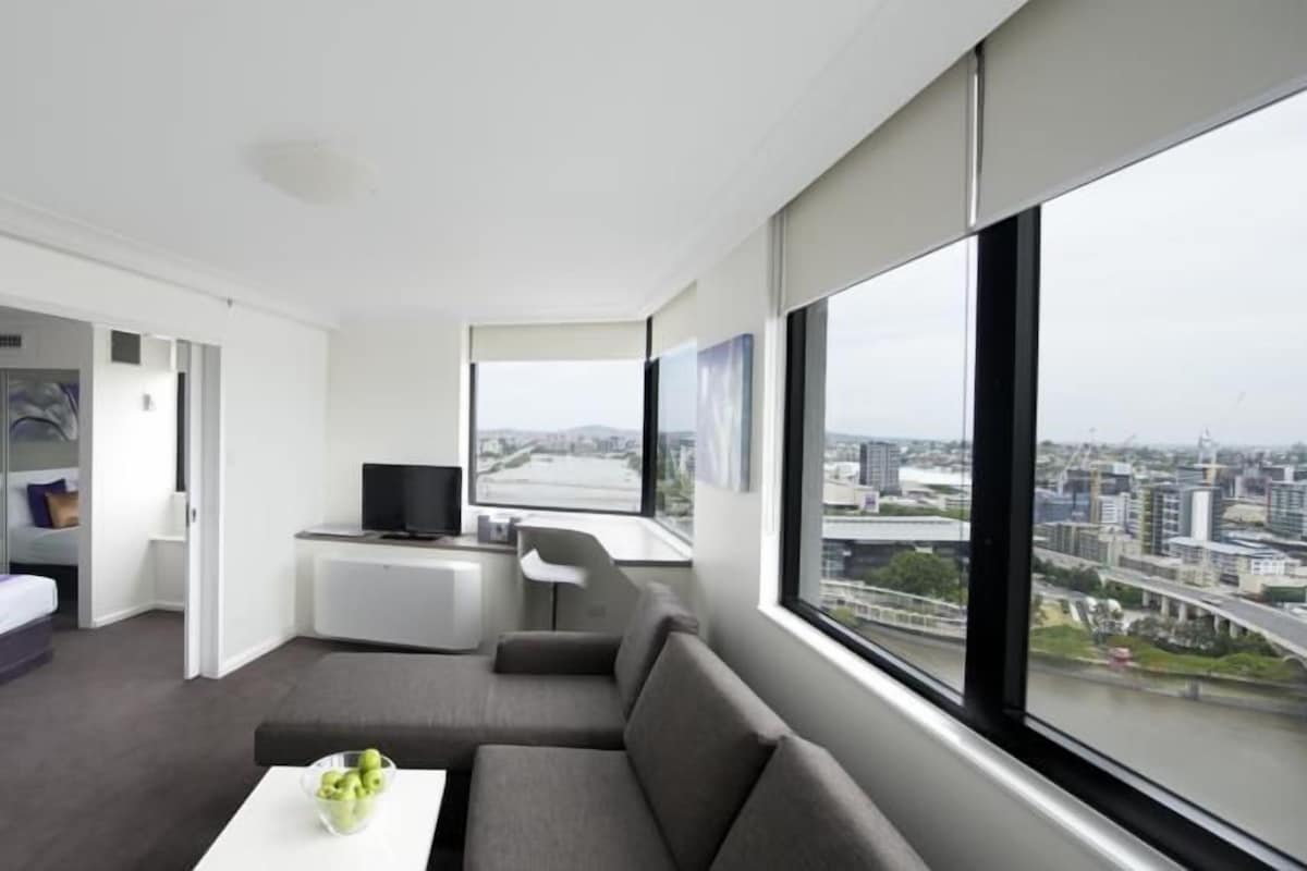 A modern, well-lit room with a large window offering a city view, a grey sofa, a small table with a bowl, and a TV on a stand.
