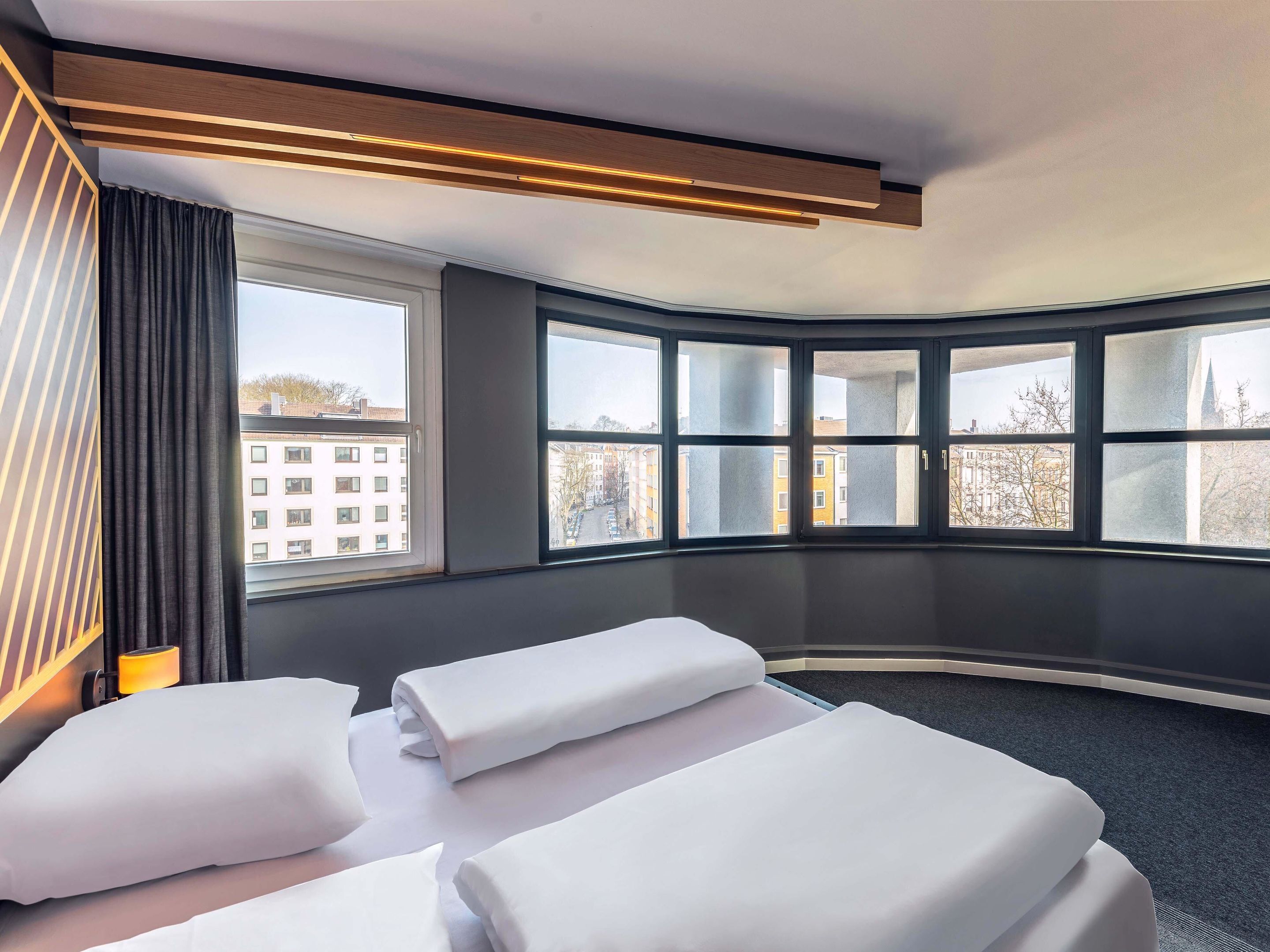 Et hotelværelse med to senge, et stort vindue med udsigt til bygninger, og et moderne loftdesign.