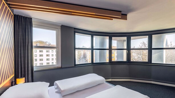 Een hotelkamer met twee bedden, een groot raam met uitzicht op gebouwen, en een modern plafondontwerp.