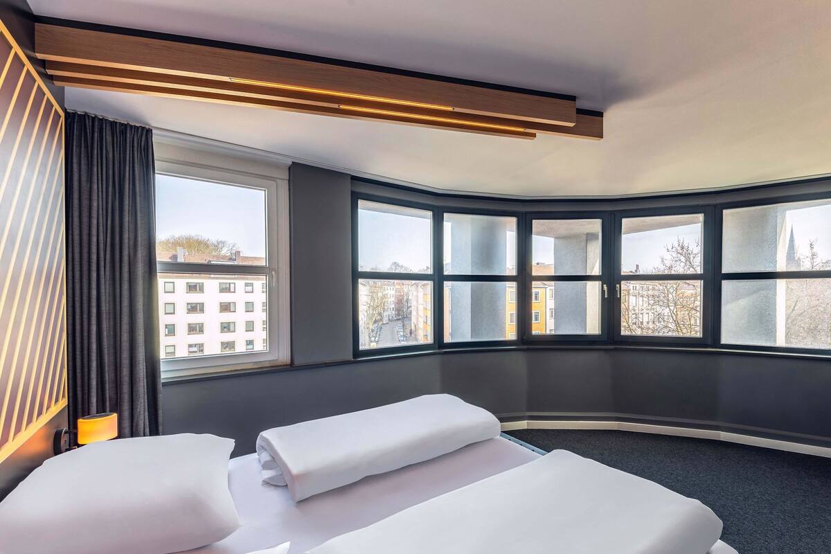 Une chambre d’hôtel avec deux lits, une grande fenêtre donnant sur des immeubles et un design moderne au niveau du plafond.