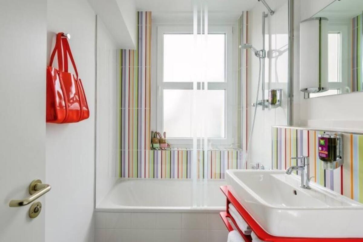 Une salle de bain avec une baignoire, un porte-serviettes rouge, un lavabo blanc et une fenêtre à rayures.