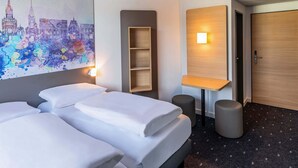 Ein Hotelzimmer mit zwei Betten, einem Stadtbildgemälde, einem Holzregal, einer Wandleuchte und einer Tür.