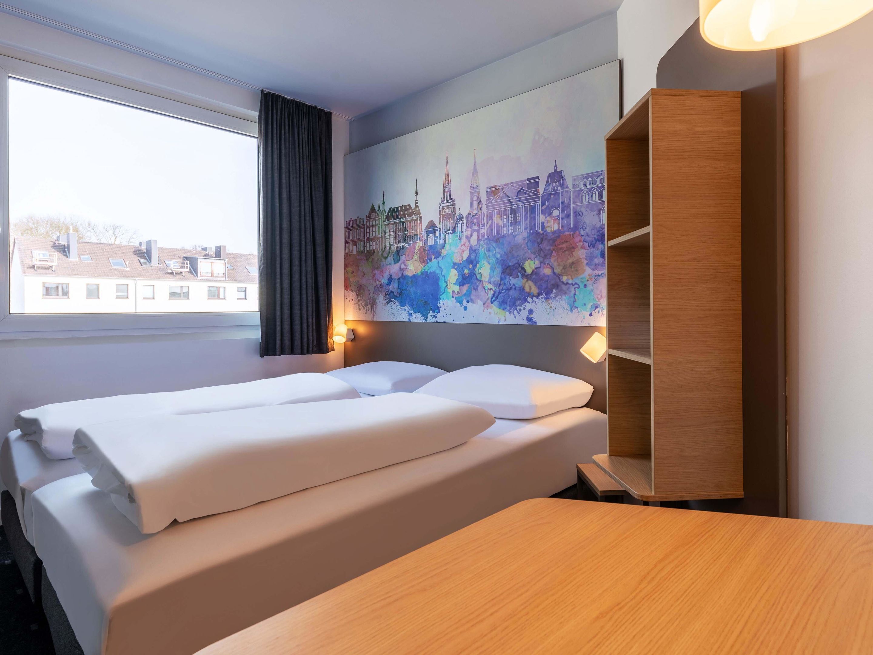 Ein Hotelzimmer mit einem Bett, einem Nachttisch aus Holz, einer Wandablage und einem Fenster mit Blick auf Gebäude.