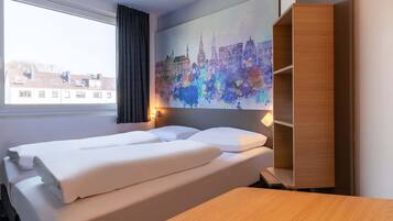 Ein Hotelzimmer mit einem Bett, einem Nachttisch aus Holz, einer Wandablage und einem Fenster mit Blick auf Gebäude.