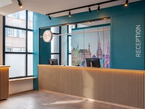 Un espace d’accueil au design moderne, comprenant un comptoir d’accueil en bois, une fresque représentant un paysage urbain et un panneau indiquant la réception.