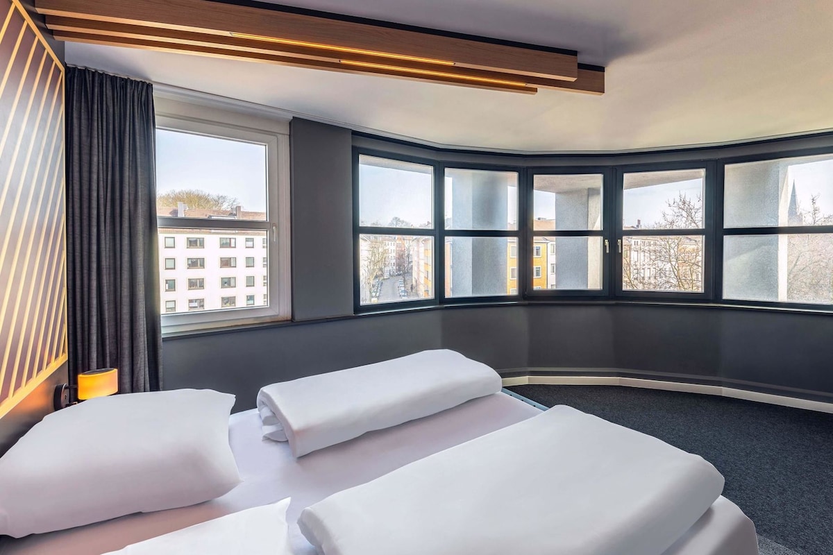 Une chambre d’hôtel avec deux lits, une grande fenêtre donnant sur des immeubles et un design moderne au niveau du plafond.