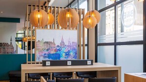 Een moderne hotel lobby met een houten receptiebalie, zwarte stoelen en een kleurrijk stadsgezichtschilderij aan de muur.