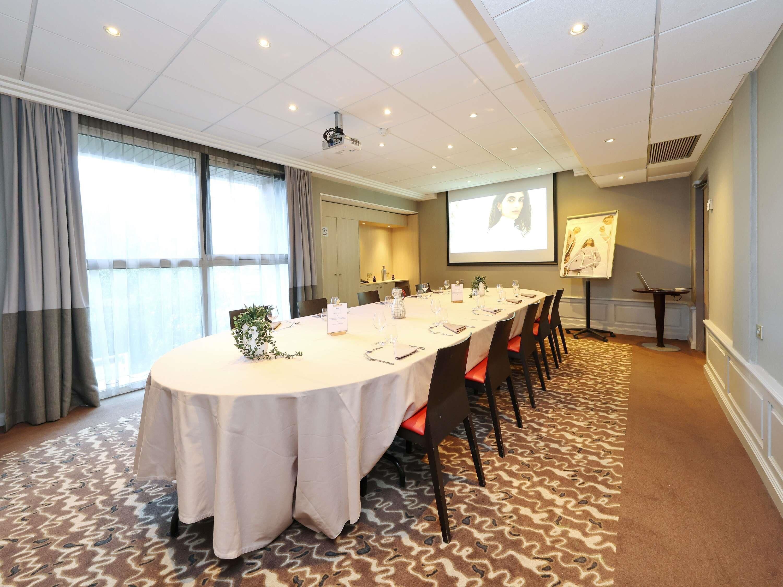 Un salón de conferencias con una larga mesa, sillas, una pantalla de proyector y un amplio ventanal.