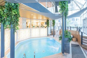 An indoor swimming pool with a wooden deck, glass walls, and green plants.