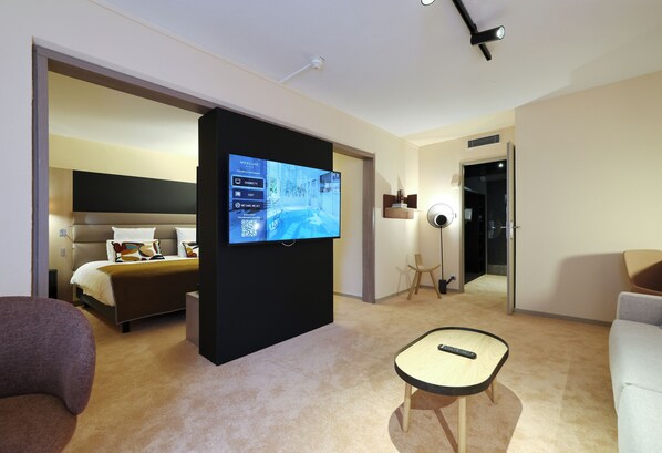 A modern hotel room with a large flat-screen TV mounted on a black panel, a bed with a headboard, a small round table with a remote control, and a light-colored carpet.
