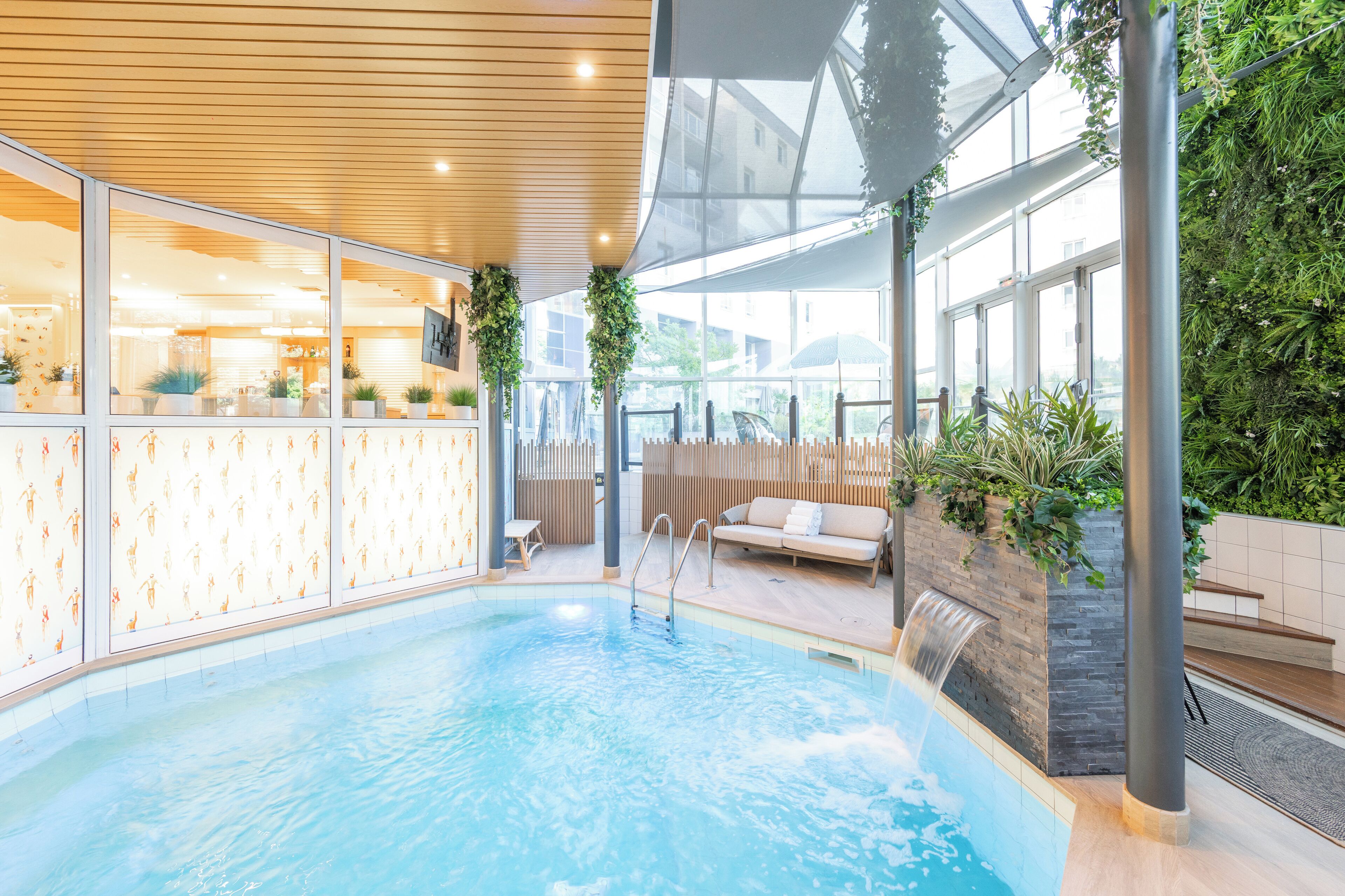An indoor swimming pool with a wooden ceiling, glass walls, and a seating area with a waterfall feature.