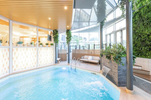 An indoor swimming pool with a wooden ceiling, glass walls, and a seating area with a waterfall feature.