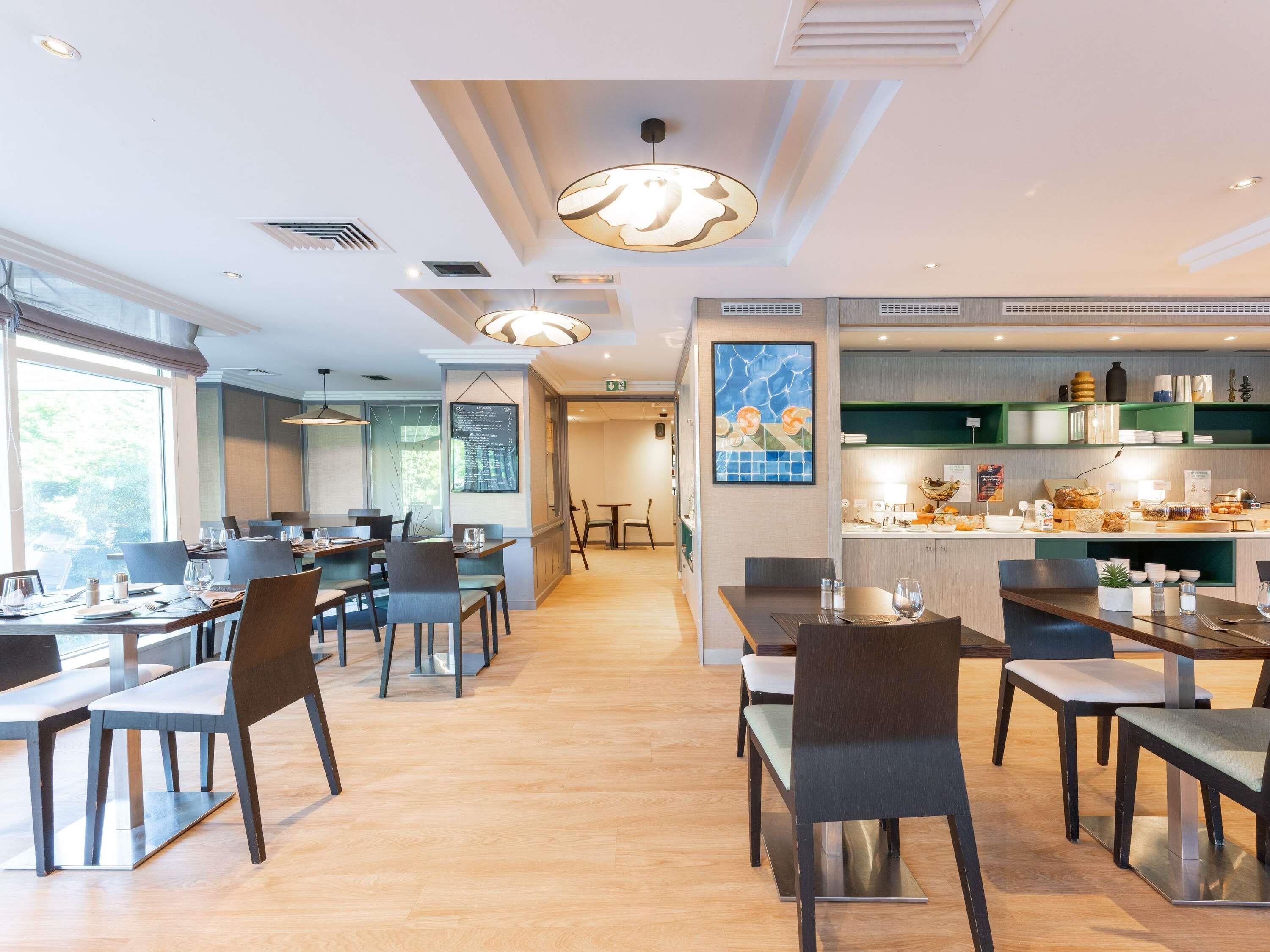 Un restaurante moderno con suelo de madera, varias mesas y sillas de comedor. Se observan luces colgantes y un menú en una pizarra en la pared.