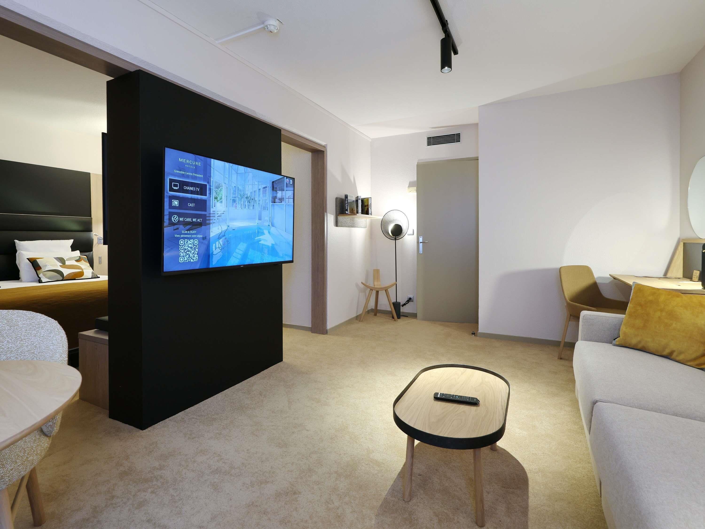 Moderni hotellihuone, jossa on taulutelevisio, sänky, sohva, pieni pöytä ja lattiatuuletin.