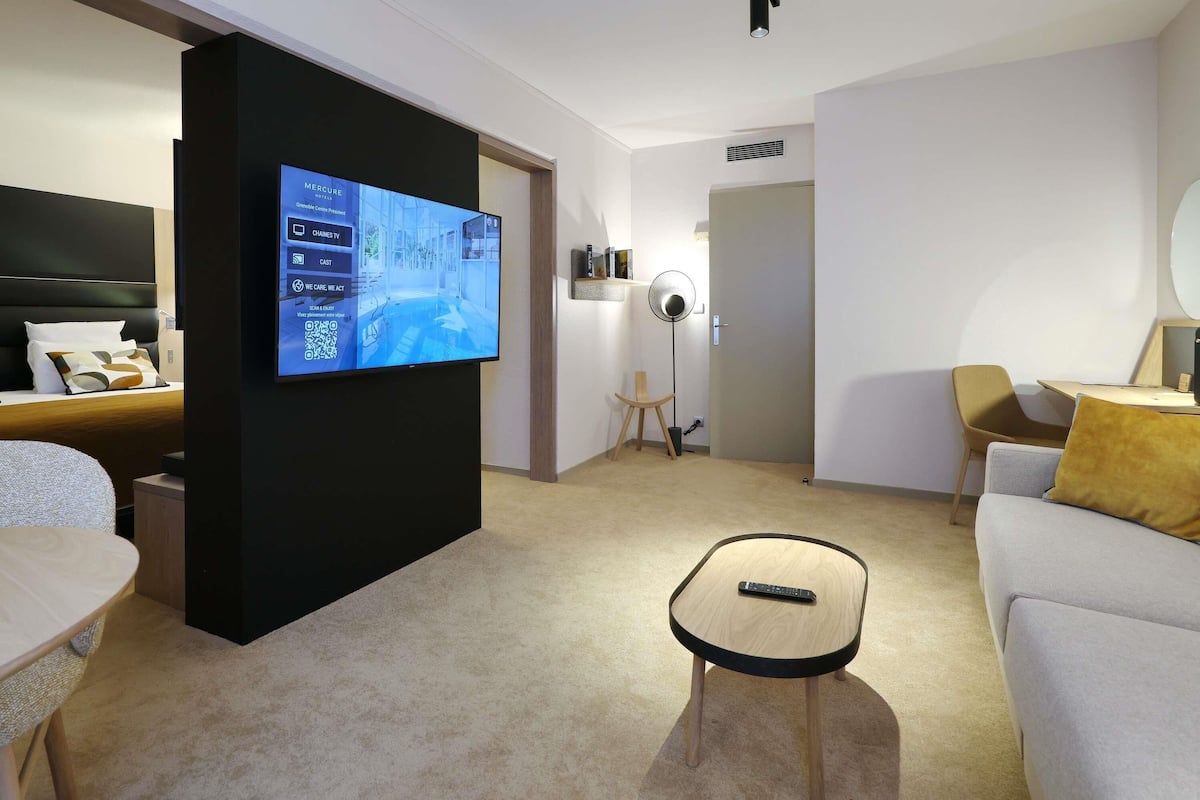Moderni hotellihuone, jossa on taulutelevisio, sänky, sohva, pieni pöytä ja lattiatuuletin.