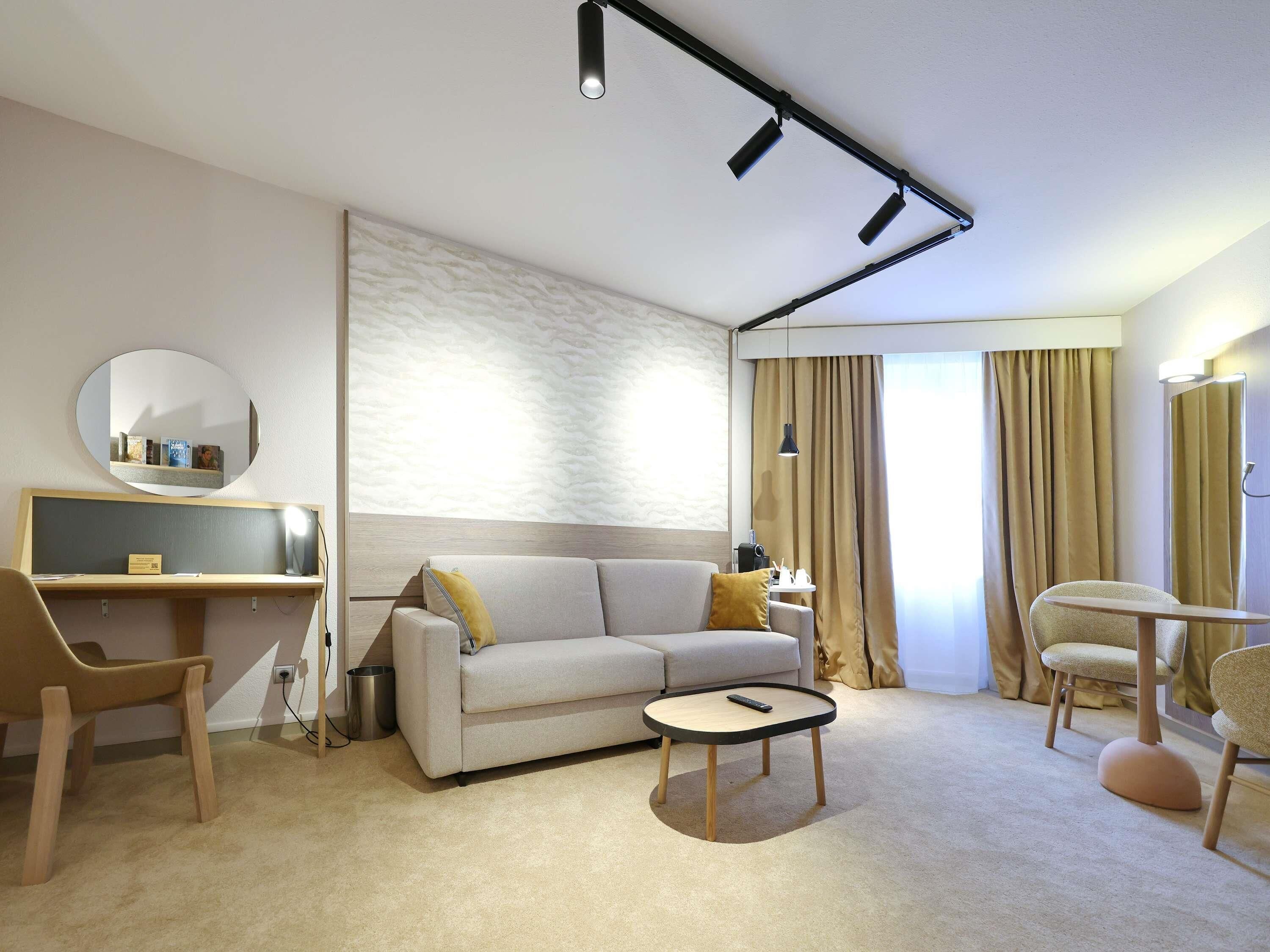 A modern hotel room with a sofa, a small round table, a desk with a chair, a mirror, and a large window with curtains.