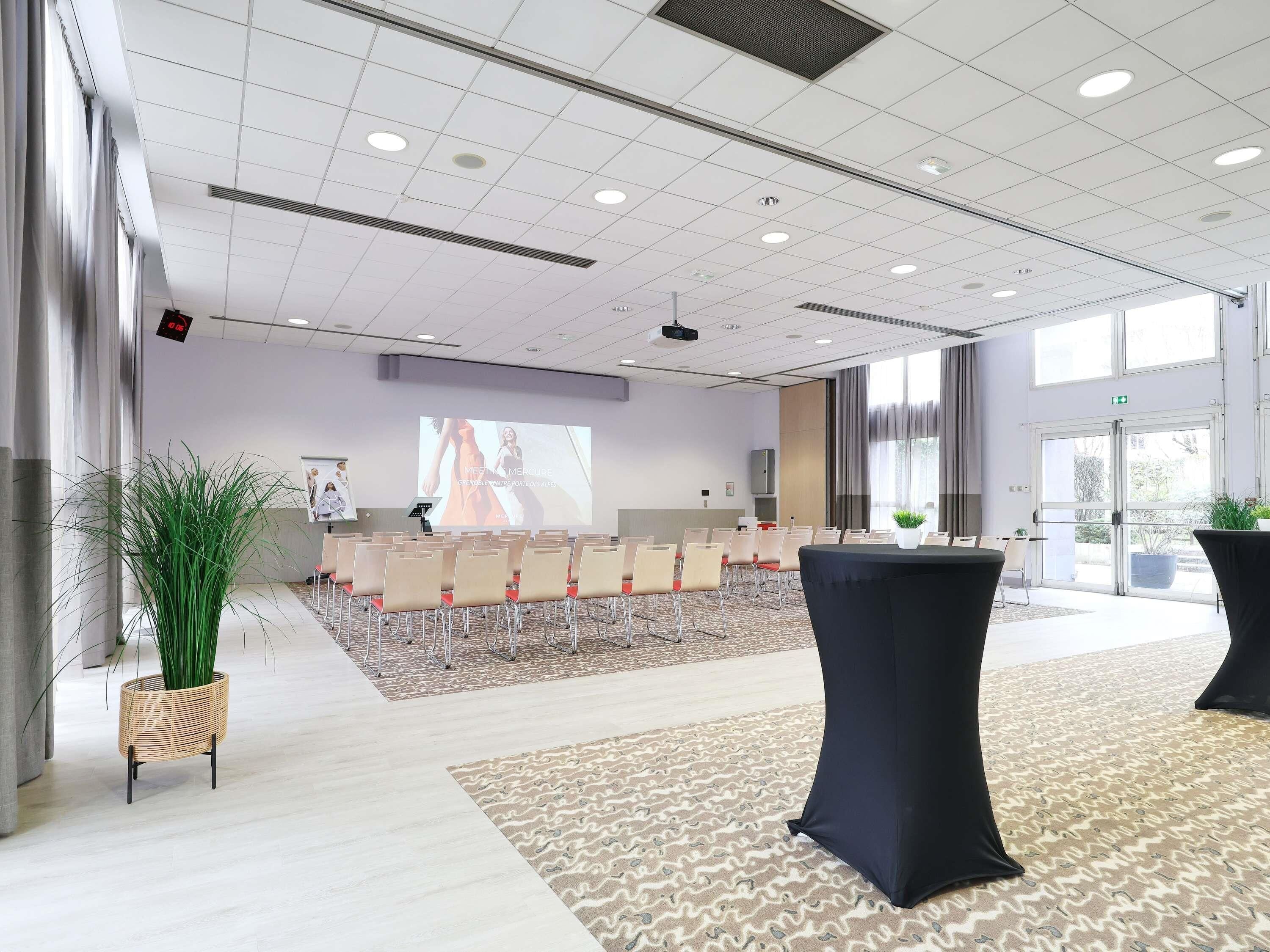 A conference room with a projector screen, rows of chairs, a large potted plant, and a patterned carpet.