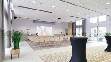A conference room with a projector screen, rows of chairs, a large potted plant, and a patterned carpet.