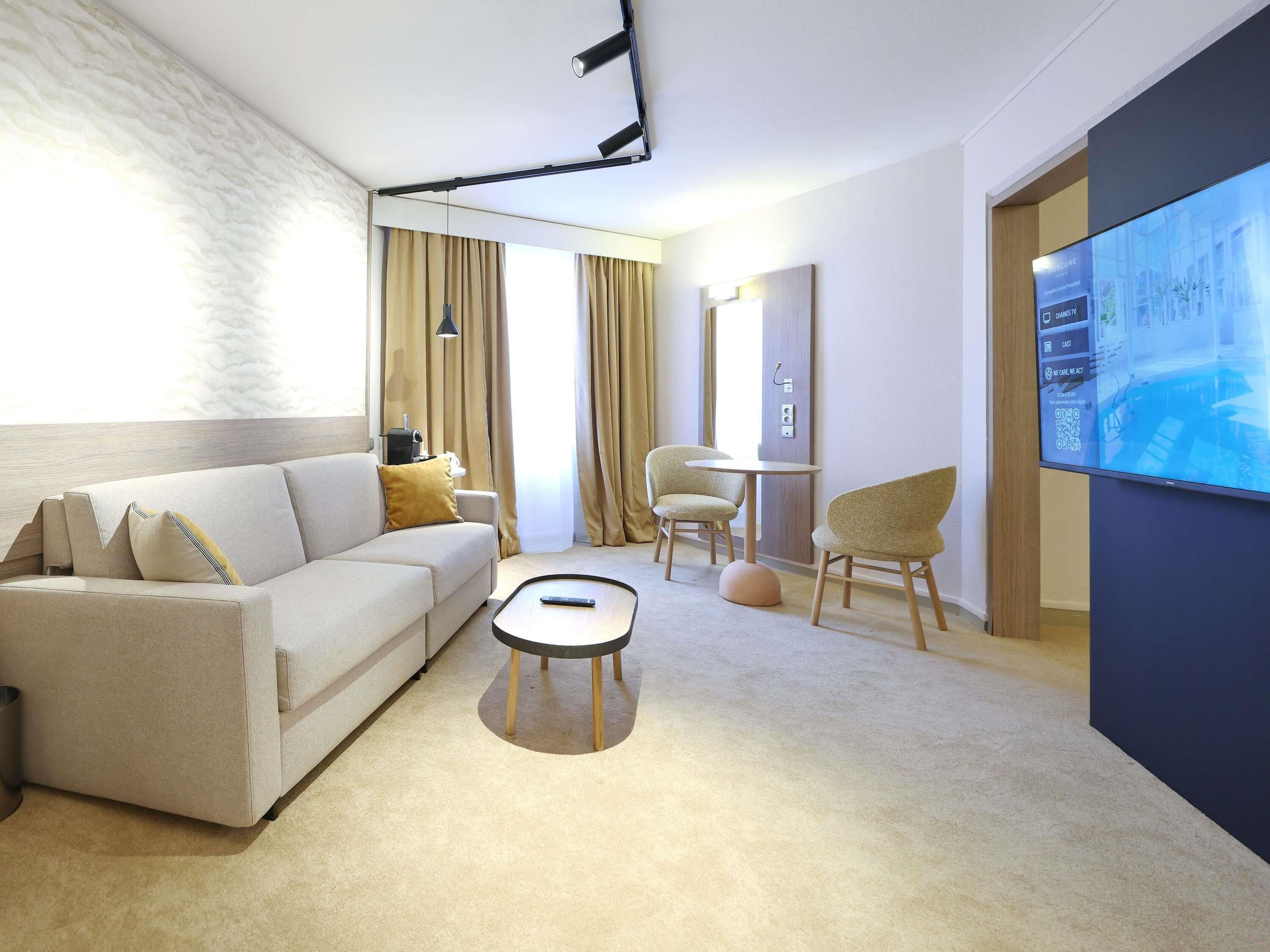 A modern hotel room with a sofa, a small round table, a chair, a TV, and a large window with curtains.