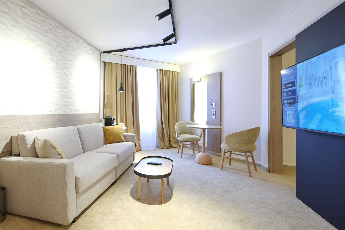 Une chambre d’hôtel moderne, équipée d’un canapé, d’une petite table ronde, d’une chaise, d’un téléviseur et d’une grande fenêtre avec des rideaux.