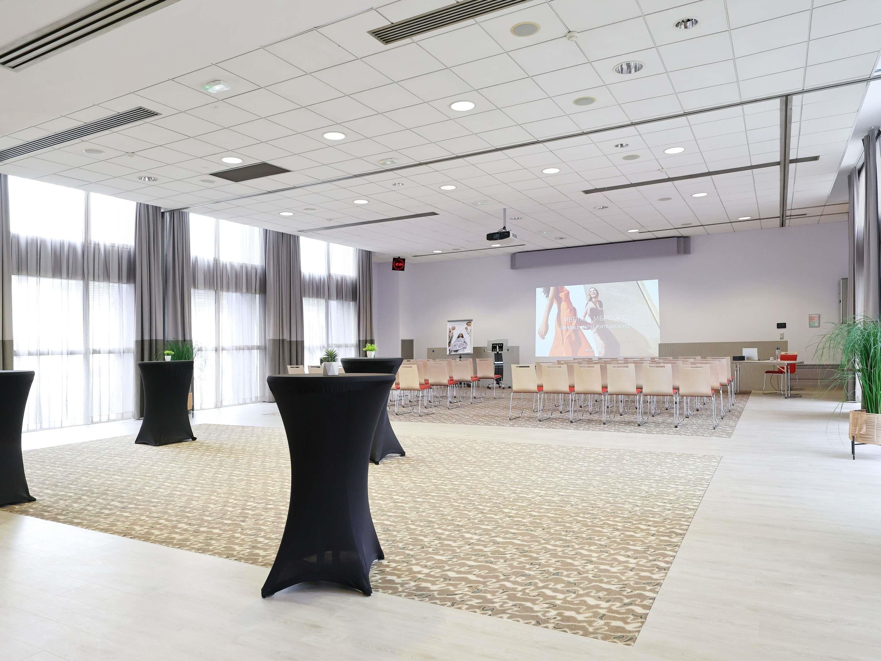 Une salle de conférence spacieuse, dotée d’un grand écran, de plusieurs tables et de chaises disposées pour les présentations.