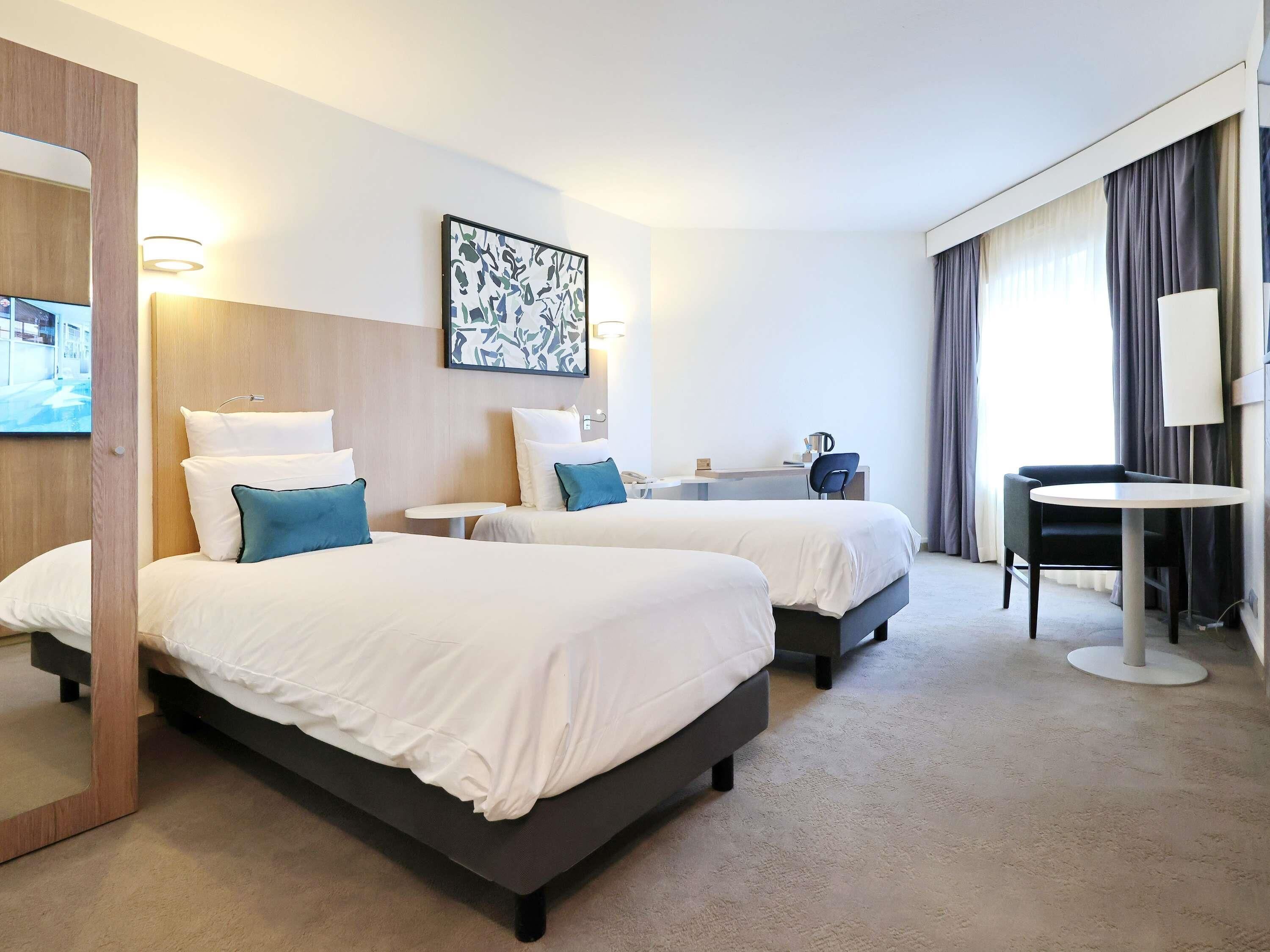 Hotellihuone, jossa on kaksi sänkyä, työpöytä, tuoli, pieni pöytä ja suuri ikkuna, jossa on verhot.