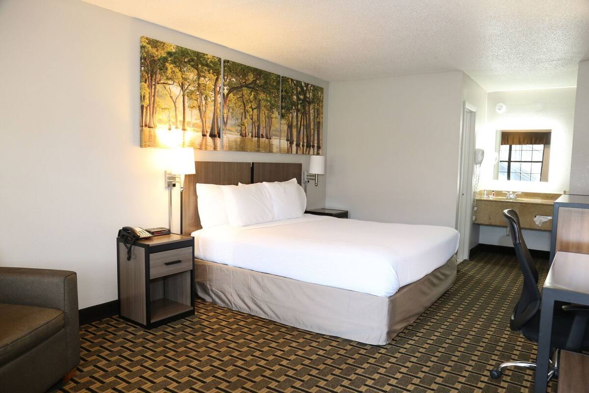 A hotel room with a bed, bedside tables, a desk, a chair, a painting on the wall, and a patterned floor.