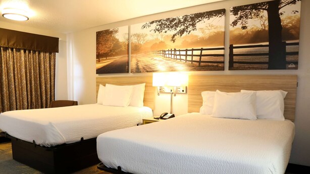 A hotel room with two beds, a wooden headboard, a nightstand with a lamp, and a large landscape painting on the wall. - Days Inn by Wyndham Monroe LA (Monroe)