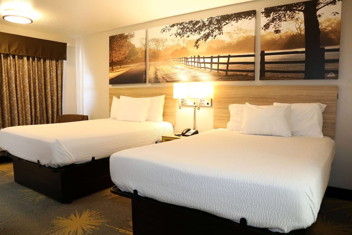 A hotel room with two beds, a wooden headboard, a nightstand with a lamp, and a large landscape painting on the wall.
