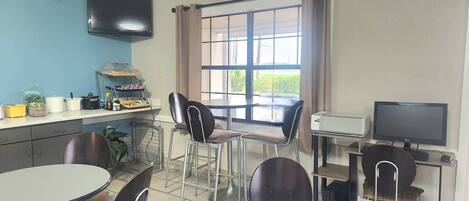 Una habitación con comedor, una encimera de cocina con electrodomésticos, televisor y una ventana con cortinas.