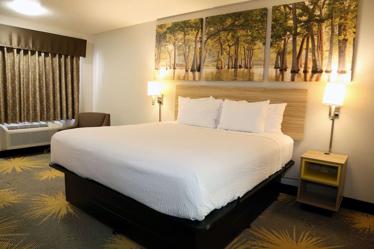 A hotel room with a large bed, two bedside lamps, a headboard, a wall-mounted air conditioner, and a window with curtains.